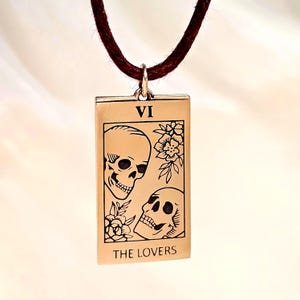 bronze-gold tone finish. Its design resembles a Tarot card, marked with the Roman numeral “VI” at the top, representing The Lovers. The artwork features two skulls facing each other, one upright and one tilted upward, with engraved floral motifs surrounding them. At the bottom of the pendant, the words “THE LOVERS” are inscribed. The pendant hangs from a small metal jump ring attached to the cord.