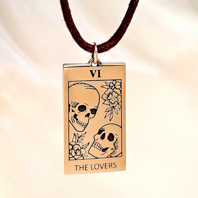 bronze-gold tone finish. Its design resembles a Tarot card, marked with the Roman numeral “VI” at the top, representing The Lovers. The artwork features two skulls facing each other, one upright and one tilted upward, with engraved floral motifs surrounding them. At the bottom of the pendant, the words “THE LOVERS” are inscribed. The pendant hangs from a small metal jump ring attached to the cord.