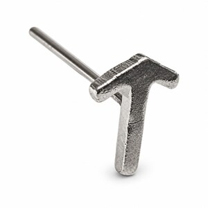 Può includere: Un orecchino a lobo in metallo color argento a forma di lettera maiuscola T. La lettera presenta una superficie testurizzata e leggermente irregolare, fissata a un perno metallico dritto e liscio che si estende dal retro del design.