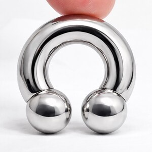 May include: A shiny, silver-colored horseshoe-shaped body jewelry piece. The jewelry has a curved bar with two round, polished balls at the ends. The piece is held by a finger.