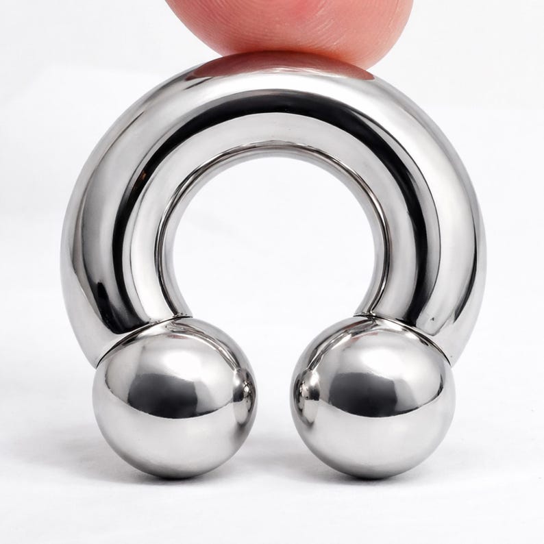 May include: A shiny, silver-colored horseshoe-shaped body jewelry piece. The jewelry has a curved bar with two round, polished balls at the ends. The piece is held by a finger.