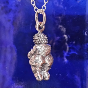 Puede incluir: Un collar con colgante de plata que presenta una figura estilizada de la Venus de Willendorf. El colgante est&aacute; suspendido de una delicada cadena de plata. La figura tiene una cabeza y un cuerpo detallados, con el n&uacute;mero "92" grabado en la parte posterior.