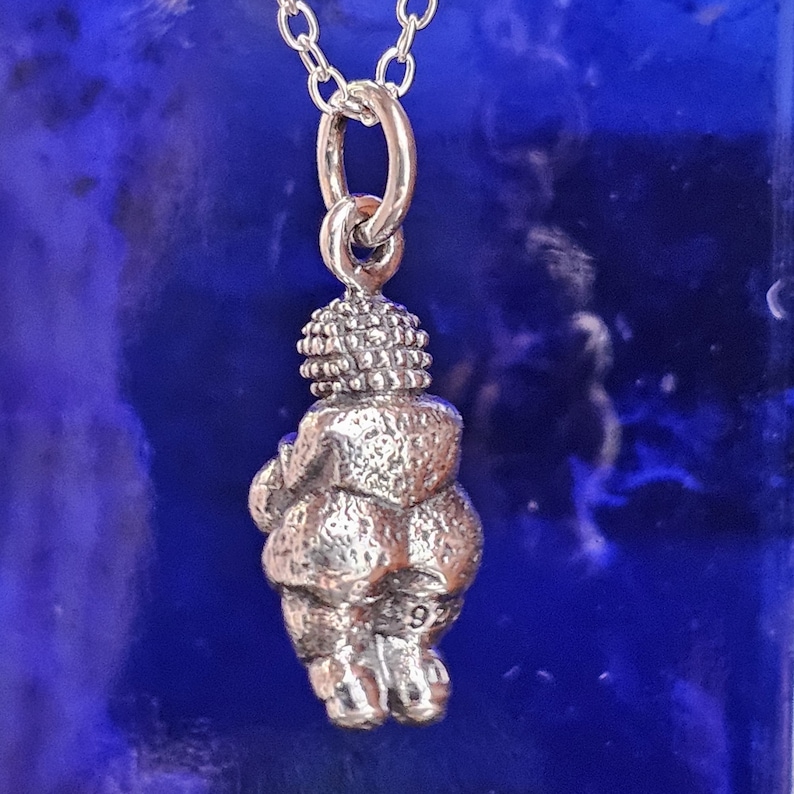 Puede incluir: Un collar con colgante de plata que presenta una figura estilizada de la Venus de Willendorf. El colgante est&aacute; suspendido de una delicada cadena de plata. La figura tiene una cabeza y un cuerpo detallados, con el n&uacute;mero "92" grabado en la parte posterior.