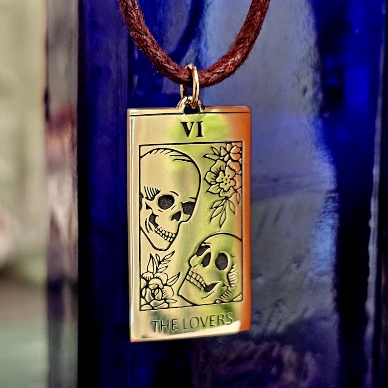 bronze-gold tone finish. Its design resembles a Tarot card, marked with the Roman numeral “VI” at the top, representing The Lovers. The artwork features two skulls facing each other, one upright and one tilted upward, with engraved floral motifs surrounding them. At the bottom of the pendant, the words “THE LOVERS” are inscribed. The pendant hangs from a small metal jump ring attached to the cord.