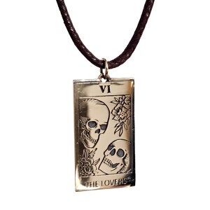 bronze-gold tone finish. Its design resembles a Tarot card, marked with the Roman numeral “VI” at the top, representing The Lovers. The artwork features two skulls facing each other, one upright and one tilted upward, with engraved floral motifs surrounding them. At the bottom of the pendant, the words “THE LOVERS” are inscribed. The pendant hangs from a small metal jump ring attached to the cord.