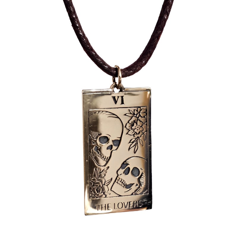 bronze-gold tone finish. Its design resembles a Tarot card, marked with the Roman numeral “VI” at the top, representing The Lovers. The artwork features two skulls facing each other, one upright and one tilted upward, with engraved floral motifs surrounding them. At the bottom of the pendant, the words “THE LOVERS” are inscribed. The pendant hangs from a small metal jump ring attached to the cord.