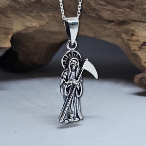Pu&ograve; includere: Collana con ciondolo in argento che presenta una rappresentazione dettagliata della Morte che tiene una falce. Il ciondolo &egrave; su una catena d'argento.