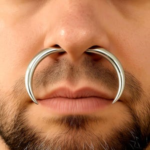 May include: A large, silver septum piercing in a horseshoe shape. The piercing is worn in the nose and curves around the nostrils. The person in the image has a beard and moustache.