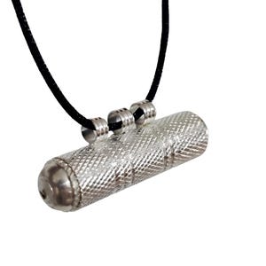 May include: Silver cylindrical pendant with a textured surface, hanging from a black cord. The pendant has rounded ends and is attached to the cord with three silver rings. The background is white.