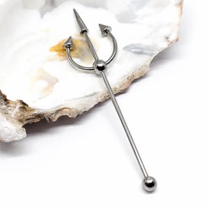 May include: A silver-colored industrial barbell featuring a trident design. The barbell has a long, straight bar with a ball at one end and a trident at the other. The trident has three points and a central spear.