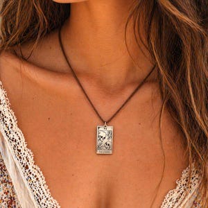May include: A silver pendant necklace featuring a rectangular charm with a skull design and the words "THE LOVERS". The pendant hangs from a brown cord necklace. The tarot card design is detailed and intricate.