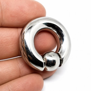 May include: A polished silver-toned captive bead ring featuring a thick, circular hoop with a smooth, reflective surface and a single spherical bead secured within the opening, creating a continuous, rounded metallic jewellery piece.