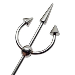 May include: A silver-colored body piercing jewelry piece in the shape of a trident. The jewelry has a central spike and two curved arms, each ending in a conical point. The design is symmetrical and has a polished finish.