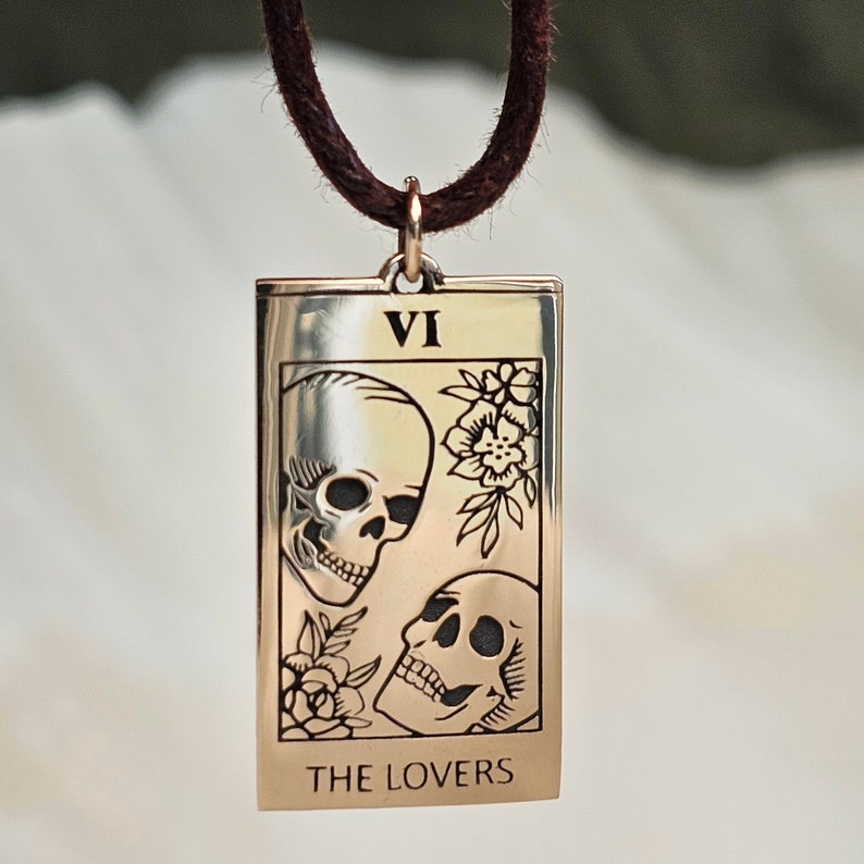 May include: A gold-colored rectangular pendant necklace featuring the tarot card "The Lovers." The card depicts two skulls, floral accents, and the Roman numeral VI. The pendant hangs from a brown cord.