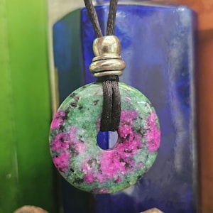 May include: A green and pink gemstone pendant with a black cord necklace. The pendant is round and has a hole in the centre.
