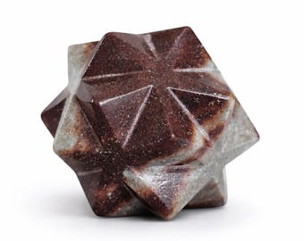 May include: A multi-faceted geometric stone sculpture featuring a star-like shape with sharp, pointed edges. The object displays a marbled texture with deep reddish-brown and muted grey color variations throughout its polished, solid surface.
