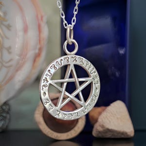 May include: Silver pendant necklace with a pentagram design and a circle of runes around the edge. The pendant is hanging from a silver chain.