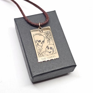 May include: A gold-colored pendant necklace featuring a tarot card design with two skulls and floral accents. The card reads "THE LOVERS" and "VI". The pendant hangs from a brown cord and rests on a black box.