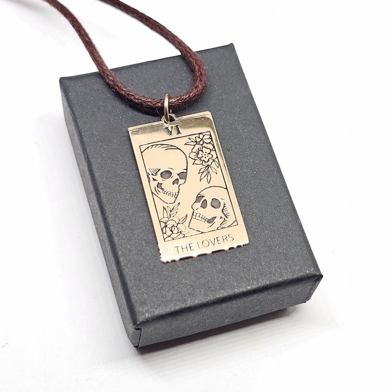 May include: A gold-colored pendant necklace featuring a tarot card design with two skulls and floral accents. The card reads "THE LOVERS" and "VI". The pendant hangs from a brown cord and rests on a black box.