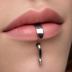 May include: Close-up of a person's lips with a silver lip piercing and a silver lip ring. The lip piercing is a flat, curved bar that sits on the lower lip. The lip ring is a curved hoop with a small ball at the end.