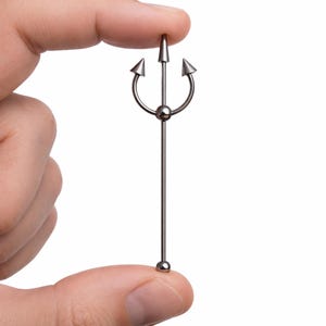 May include: A silver-colored industrial barbell body piercing jewelry. The jewelry features a trident design at the top and a small ball at the bottom. The barbell is held between two fingers against a white background.