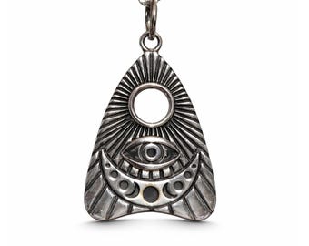 Könnte beinhalten: Ein silberfarbener Metallanhänger in Form einer Planchette, der an einer passenden Kette hängt. Der Anhänger zeigt ein geprägtes Design mit einem zentralen Auge, einem Halbmond mit kreisförmigen Phasen und strahlenförmigen Linien um eine Aussparung.