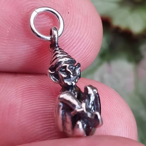 May include: A silver charm featuring a detailed sculpture of a gnome wearing a pointed hat. The gnome is sitting with its hands clasped in front of it.