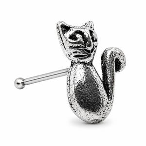 May include: A silver-colored nose stud in the shape of a cat. The cat is depicted in a sitting position with its tail curled up. The nose stud has a straight post with a small ball on the end.