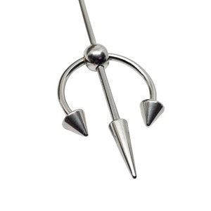 May include: A silver-colored industrial barbell body piercing. The jewelry features a central, long, conical spike and two curved spikes on either side, resembling a trident. The design is simple and metallic, set against a white background.