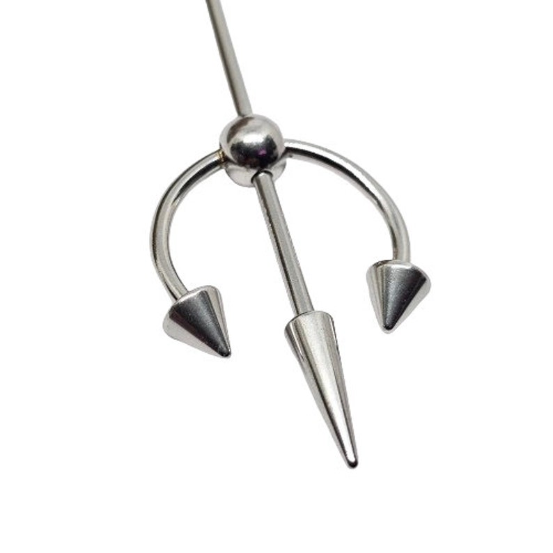 May include: A silver-colored industrial barbell body piercing. The jewelry features a central, long, conical spike and two curved spikes on either side, resembling a trident. The design is simple and metallic, set against a white background.