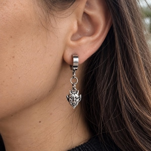 May include: A silver-toned hoop earring featuring a small, dangling charm shaped like a horned devil face. The earring consists of a smooth circular hoop connected by a small jump ring to the detailed, metallic pendant with pointed ears.