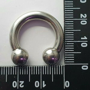 May include: A silver circular barbell with two ball ends. The barbell is measured against a ruler, showing it is approximately 1 inch in diameter.