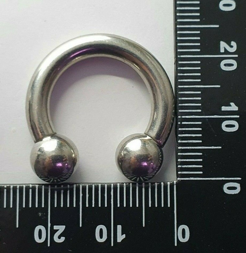 May include: A silver circular barbell with two ball ends. The barbell is measured against a ruler, showing it is approximately 1 inch in diameter.