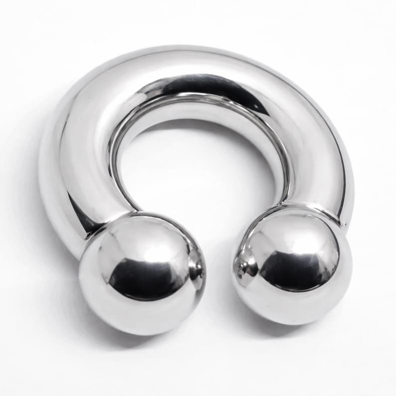 May include: A shiny, silver-colored horseshoe-shaped body jewelry piece. The jewelry has a curved bar with a smooth, rounded surface and two spherical ends. The piece is designed to be worn as a piercing.