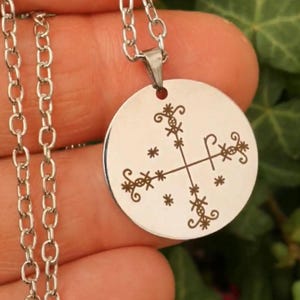 Könnte beinhalten: Eine silberne Kettenkette mit einem runden Anhänger mit einem gravierten Silbersymbol. Das Symbol ist ein Kreuz mit komplizierten Designs und kleinen Sternen.