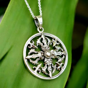 Peut inclure: Un collier pendentif en argent avec un design circulaire détaillé et des motifs complexes. Le pendentif, suspendu à une fine chaîne en argent, présente un élément central orné. Le collier est présenté sur une feuille verte.