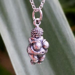 Puede incluir: Un collar con colgante de plata con una peque&ntilde;a y detallada escultura de la Venus de Willendorf. El colgante est&aacute; suspendido de una delicada cadena de plata. La escultura representa una figura femenina estilizada y curvil&iacute;nea.