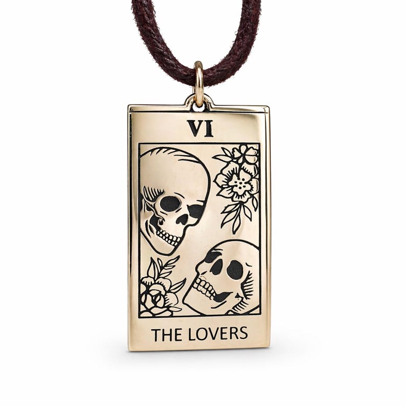 May include: Gold-colored pendant necklace featuring a tarot card design. The rectangular pendant displays two skulls, floral accents, and the text "THE LOVERS" and "VI". The necklace has a brown cord.