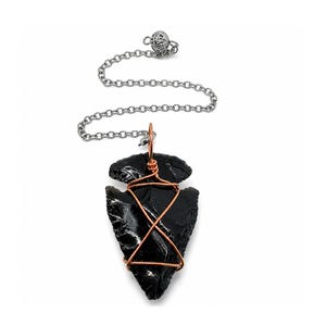 May include: A black obsidian arrowhead pendant wrapped in copper wire, suspended from a silver-toned metal chain. The arrowhead has a glossy, smooth surface with sharp, jagged edges, and the copper wire forms a geometric cage around the stone.
