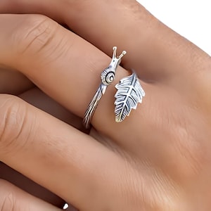 May include: A silver ring with a snail and leaf design. The snail has a detailed shell and antennae, while the leaf is intricately carved. The ring wraps around the finger, creating a unique and nature-inspired accessory.