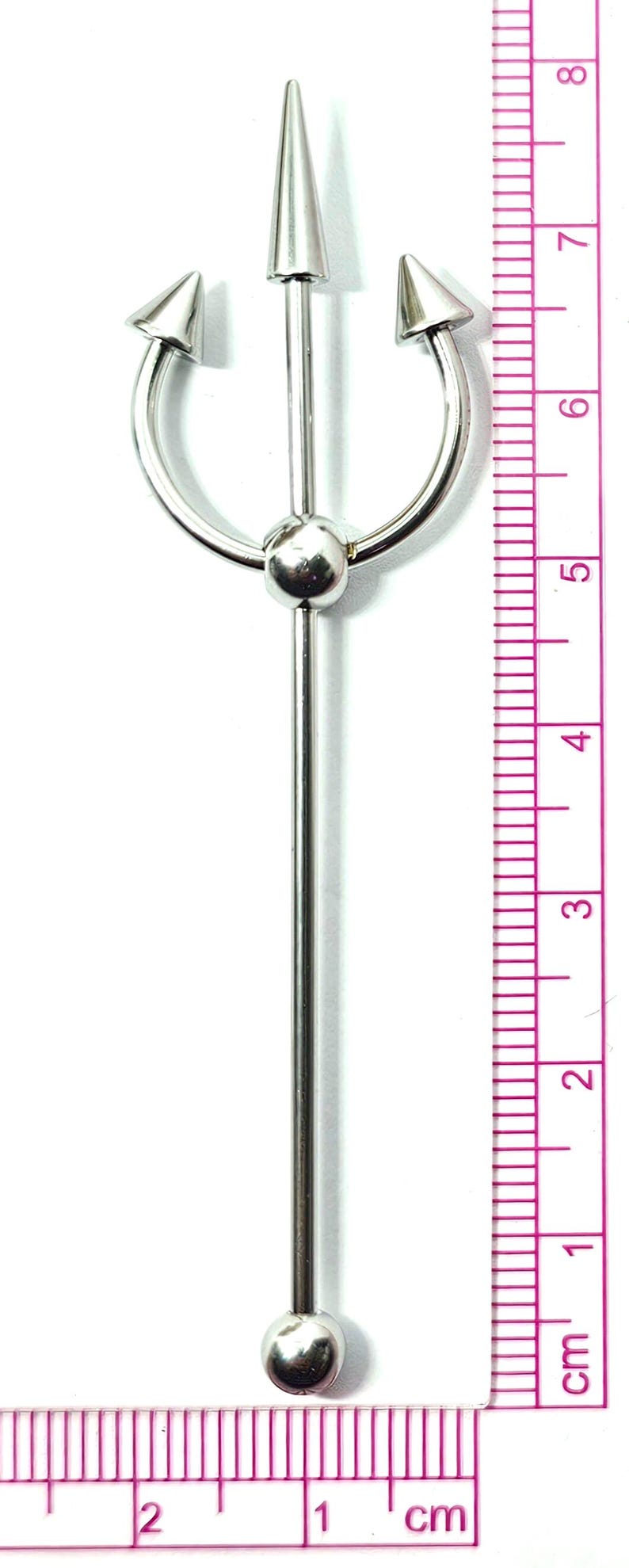 May include: A silver-colored body piercing jewelry piece. The design features a central spike, two curved spikes, and a central ball. The piece measures approximately 8 cm in length.