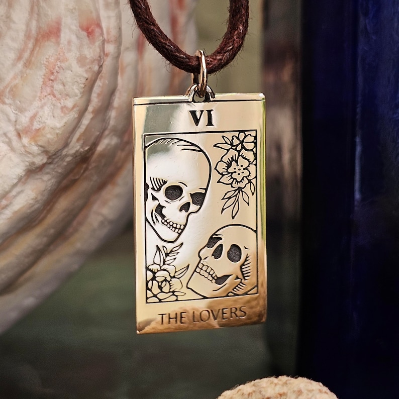 May include: A rectangular gold-colored pendant with a brown cord. The pendant features a tarot card design with two skulls, flowers, and the text "THE LOVERS" and the Roman numeral "VI".