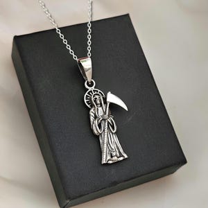 Pu&ograve; includere: Una collana d'argento con un ciondolo raffigurante una figura scheletrica che tiene una falce. La figura indossa una tunica con cappuccio e ha un'aureola sopra la testa.