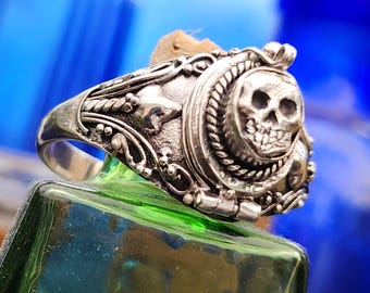 May include: A silver ring featuring a detailed skull design. The skull is the focal point, surrounded by ornate rope and floral patterns. The ring is set against a blurred background of green and blue.