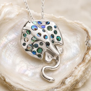 Può includere: Collana con pendente a forma di manta in argento. Il pendente presenta intarsi di conchiglia di abalone, creando un motivo colorato e strutturato. La collana è esposta su una conchiglia bianca.