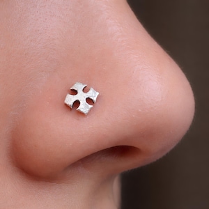 May include: A small, silver-coloured nose stud featuring a four-pointed cross shape with rounded edges. The metal surface has a textured, matte finish, and the piece is positioned flat against the skin of the side of a nose.