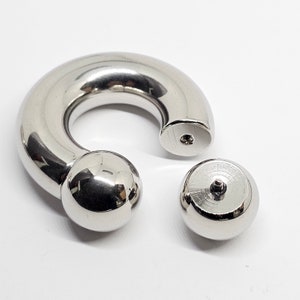 May include: A shiny, silver-colored body jewelry piece, composed of a curved barbell and two detachable ball ends. The barbell has a rounded shape with a ball at one end. The other end has a flat surface with a small hole. The detached ball is also silver.