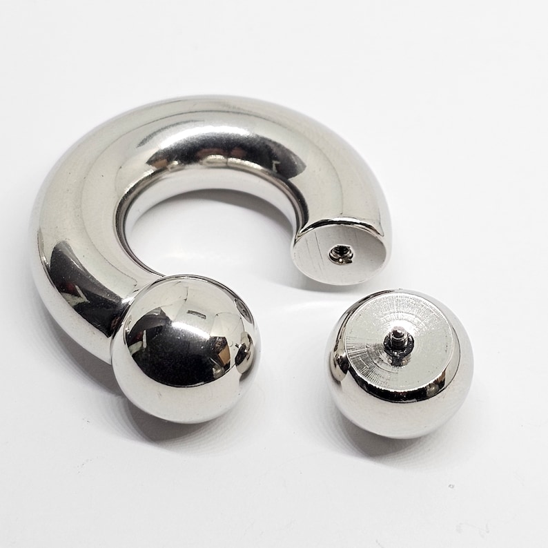 May include: A shiny, silver-colored body jewelry piece, composed of a curved barbell and two detachable ball ends. The barbell has a rounded shape with a ball at one end. The other end has a flat surface with a small hole. The detached ball is also silver.
