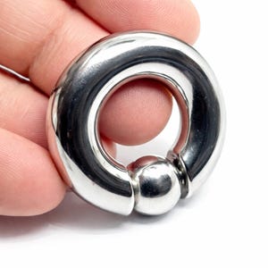 Pode incluir: Um anel circular de contas cativas feito de metal polido em tom prateado. A peça de joalharia apresenta um aro grosso e liso com uma única conta esférica mantida no lugar pela tensão do anel, criando uma superfície metálica contínua e reflexiva.