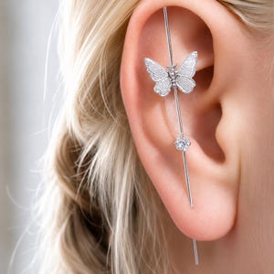 May include: A silver-toned industrial barbell earring featuring a textured butterfly charm and a single clear round gemstone. The long, straight metal bar passes through the upper ear cartilage, extending vertically along the outer ear edge.